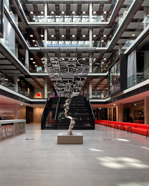 Moderner Innenbereich mit einer geschwungenen Skulptur, zentraler Treppe, roten Sofas und Kunstinstallation an der Decke.